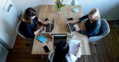Three women collaborate at a table with laptops, coffee, and notes, brainstorming UGC social media strategies.