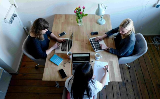 Three women collaborate at a table with laptops, coffee, and notes, brainstorming UGC social media strategies.